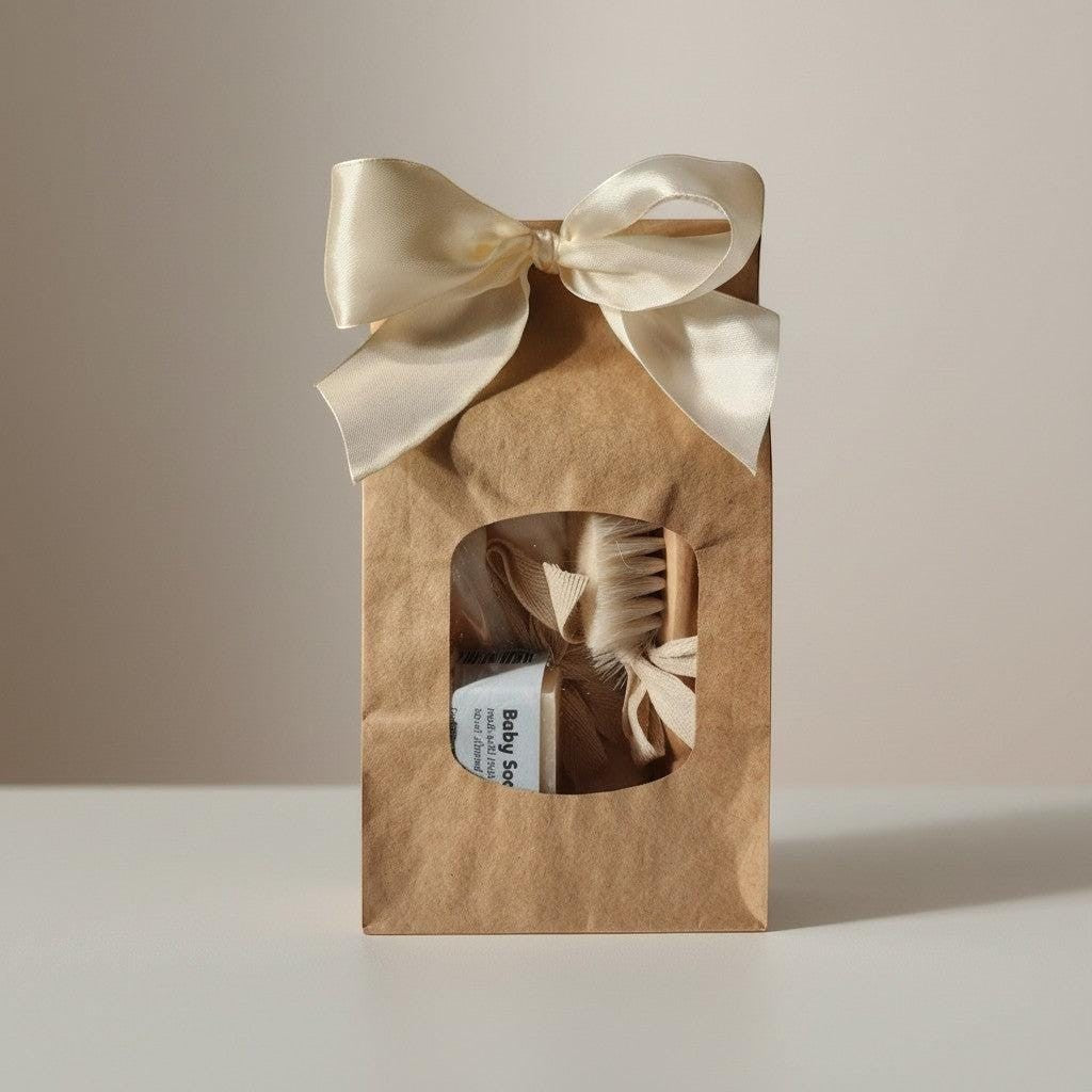 Precious Days Gifts baby bath essentials gift set in a brown paper bag with a cream ribbon bow. Includes washcloths, soap, and a brush.