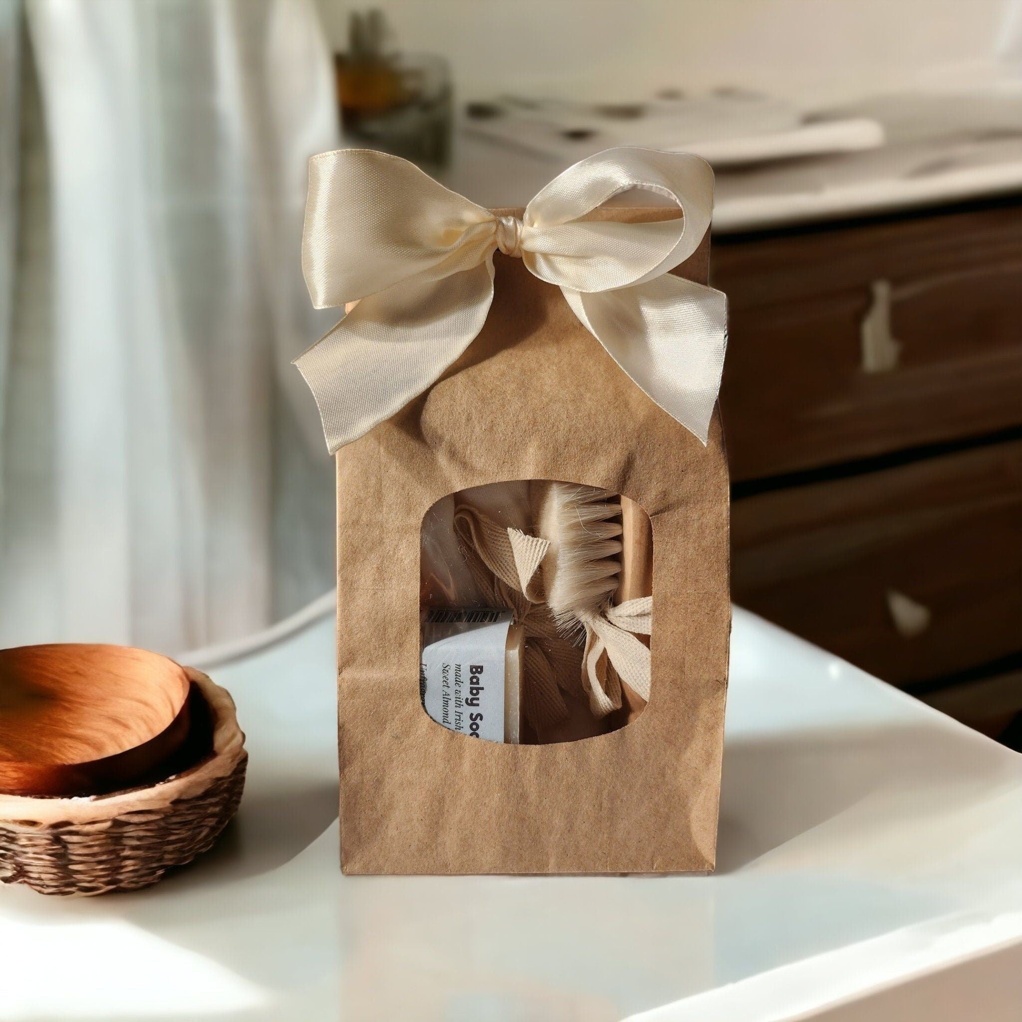 Precious Days Gifts baby bath gift set in a brown paper bag with a cream ribbon bow, includes washcloths, brush, comb, and soap.