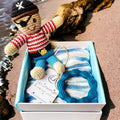 Precious Days Gifts pirate baby gift box with crochet toy, blue striped muslin, and teal teether on sand.