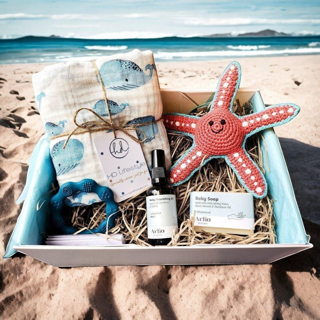 Precious Days Gifts baby gift box with whale muslin, starfish rattle, blue teether, baby oil, and soap on a beach.