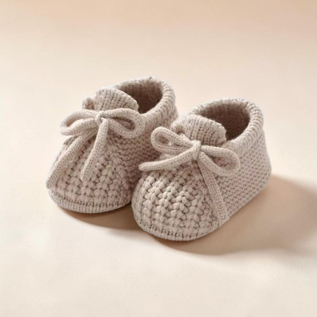 Precious Days Gifts beige cable-knit baby booties featuring a decorative front bow and soft, flexible construction.