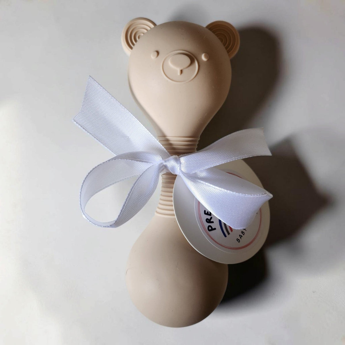 Precious Days Gifts beige silicone teddy bear rattle with a white ribbon bow for babies