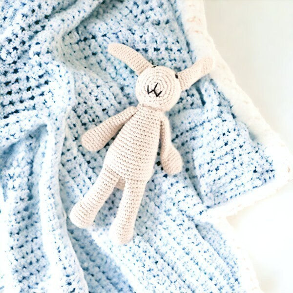 Precious Days Gifts beige crochet bunny rattle on a light blue textured blanket with white trim.