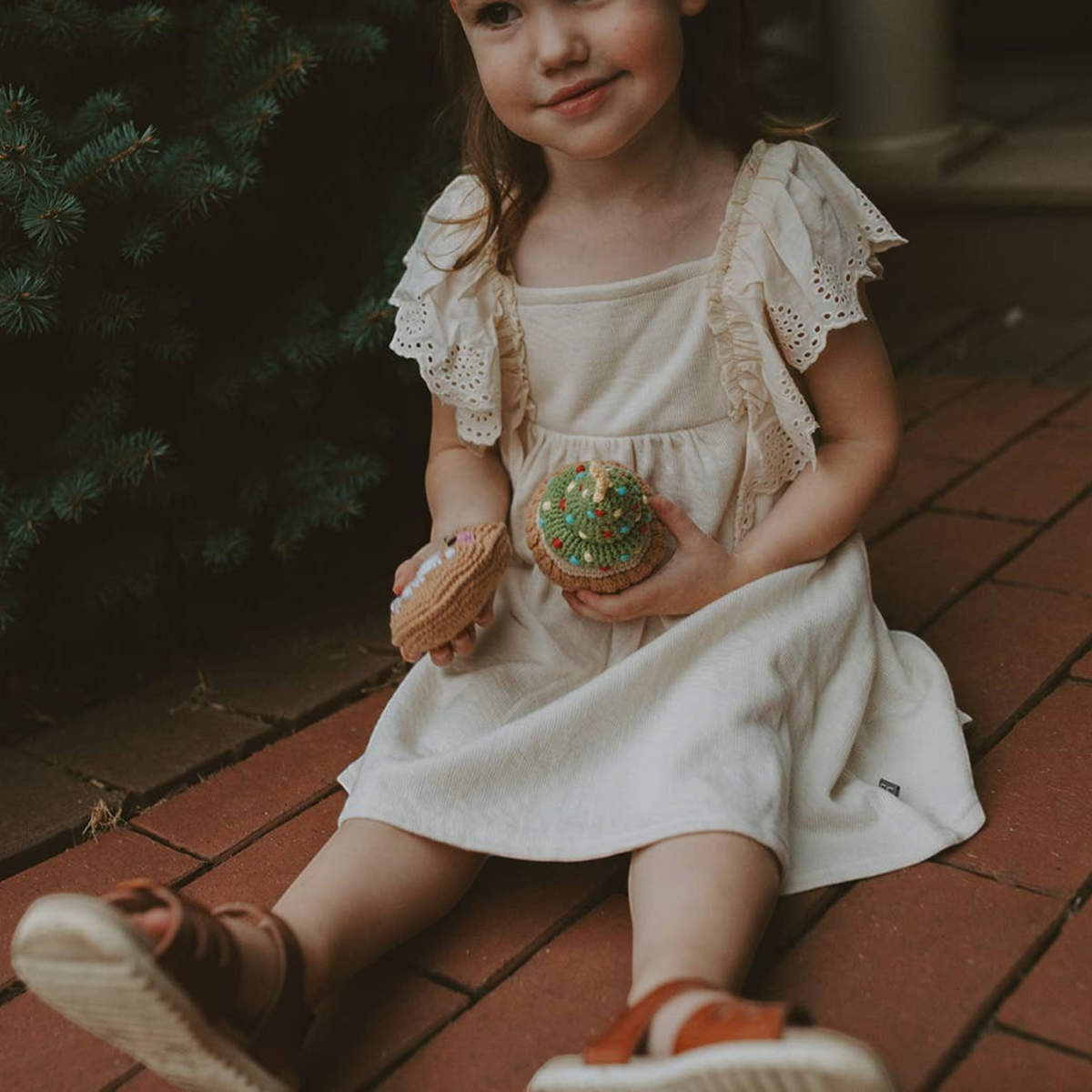 Young girl sitting on a brick porch holding Precious Days Gifts organic cotton Christmas sugar cookie and cupcake rattles