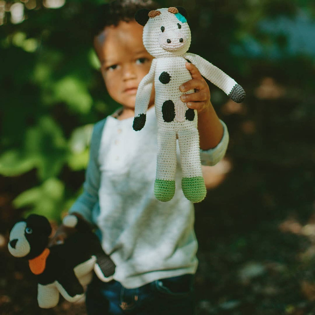 Precious Days Gifts handmade organic cotton cow plush toy with black spots and green feet, held by a child.
