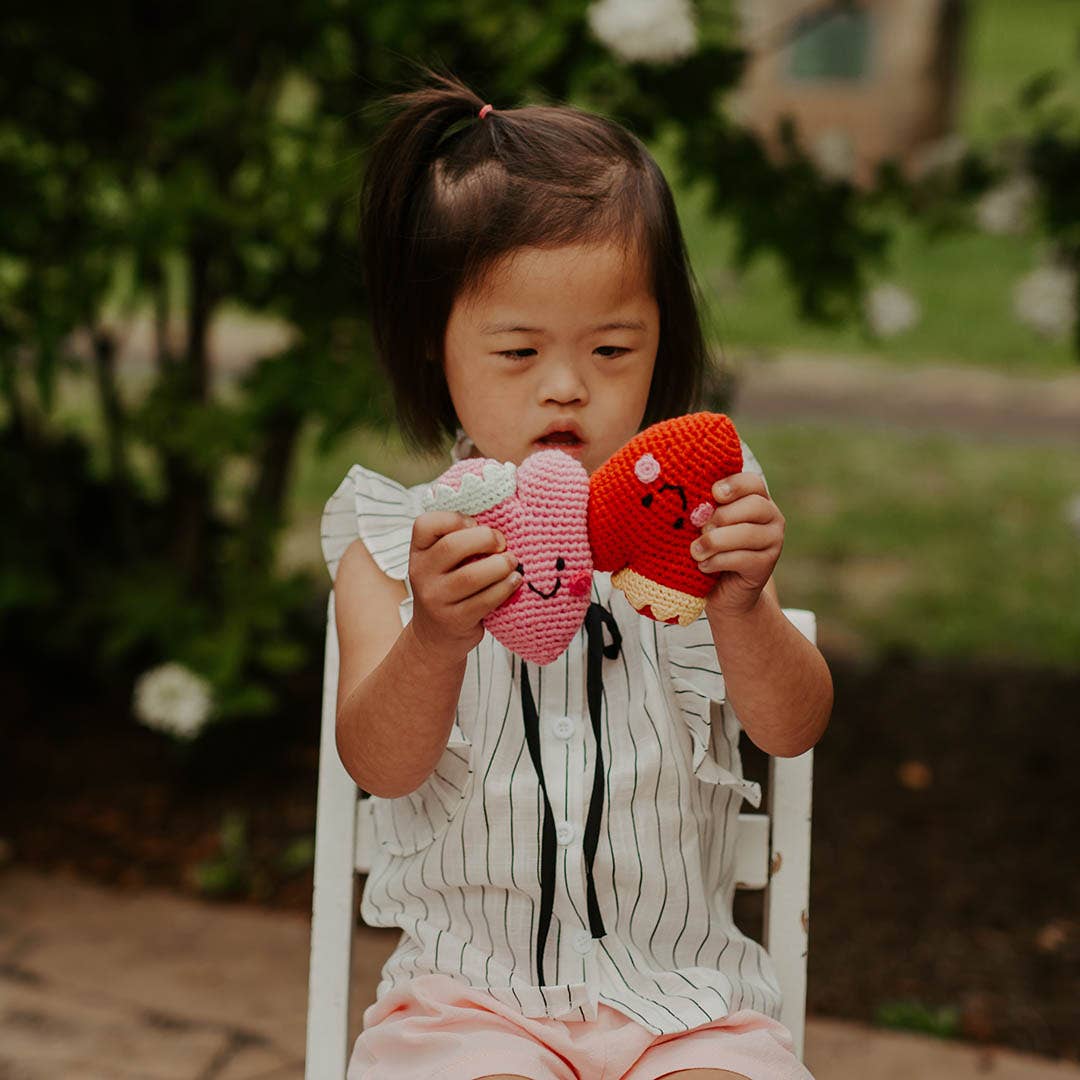 A young girl holding two Precious Days Gifts heart-shaped crochet rattles in pink and red while sitting outdoors.