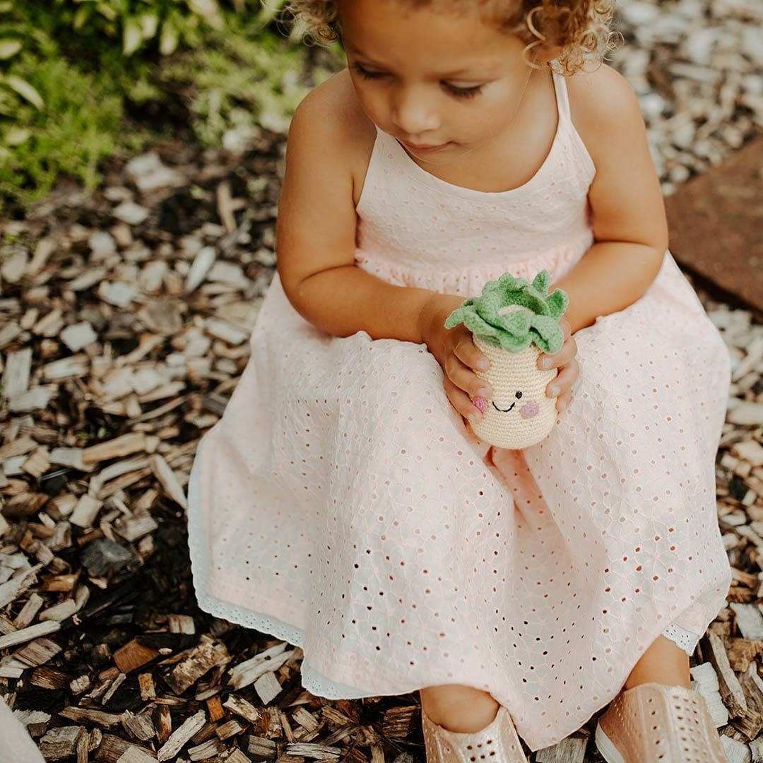 Precious Days Gifts crocheted pineapple rattle toy with green leaves and a smiling face, held by a young girl.