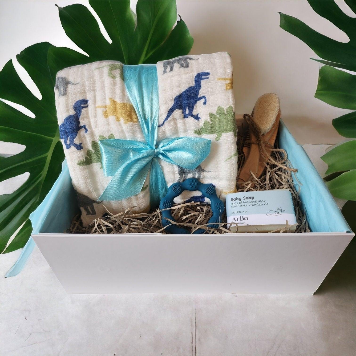 Precious Days Gifts baby gift box with a dinosaur print muslin blanket, teether, baby soap, and brush.