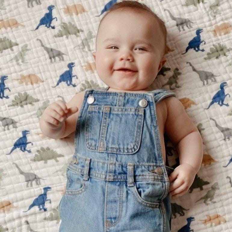 Precious Days Gifts dinosaur print 6-layer cotton muslin blanket with a smiling baby wearing denim overalls.