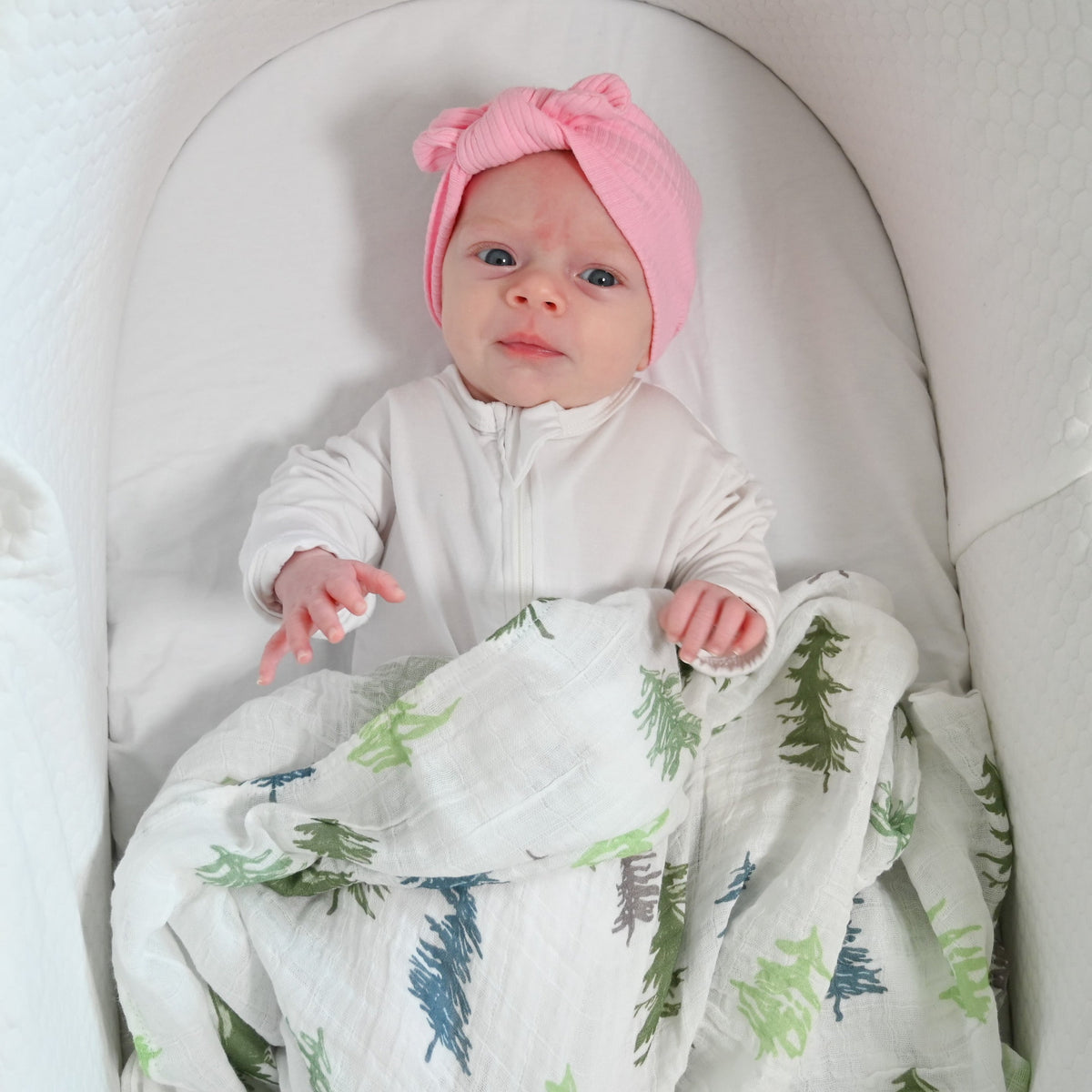 Precious Days Gifts baby wearing a pink hat, wrapped in a soft cotton muslin swaddle with a green fir tree print.
