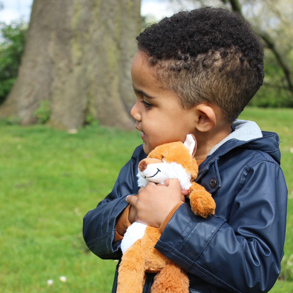 A young boy holding a soft orange and white Precious Days Gifts fox plush toy in a park setting.