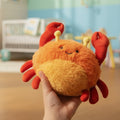 Precious Days Gifts soft orange plush crab toy with friendly expression, held in hand in a nursery setting.