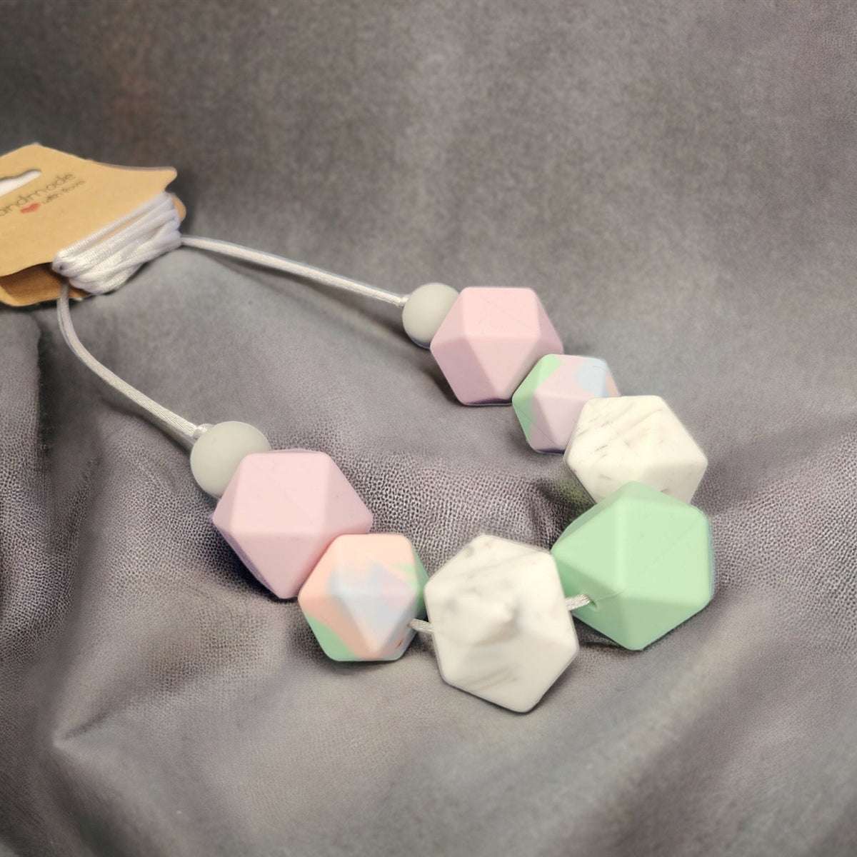 Precious Days Gifts pastel silicone bead breastfeeding necklace with geometric shapes on a grey fabric background