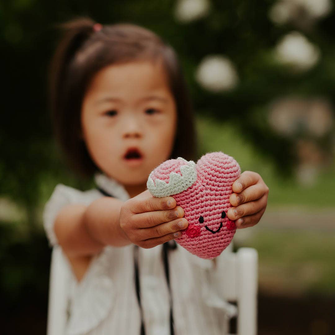 Precious Days Gifts pink heart-shaped crochet rattle held by a young girl in a garden setting