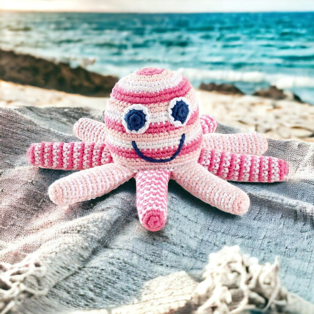 Precious Days Gifts pink and white striped crochet octopus rattle with blue eyes and a smiling mouth, on a beach towel by the sea.