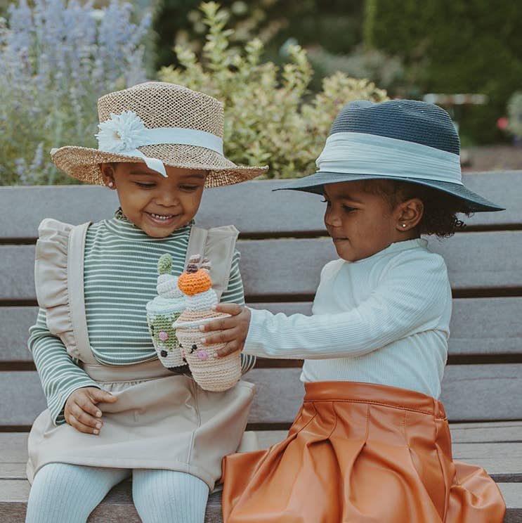 Two young girls in sun hats sitting on a bench holding a Precious Days Gifts handmade pumpkin spice latte toy rattle.