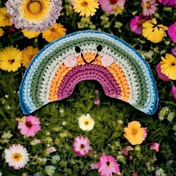 Precious Days Gifts crocheted rainbow rattle with smiling face, set amongst colorful flowers.