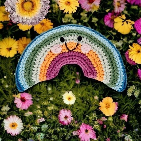 Precious Days Gifts crocheted rainbow rattle with smiling face, set amongst colorful flowers.