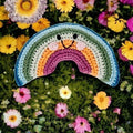 Precious Days Gifts crocheted rainbow rattle with smiling face, set amongst colorful flowers.