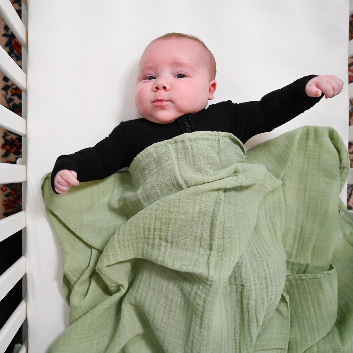 Precious Days Gifts sage green cotton muslin swaddle blanket covering a baby lying in a white crib
