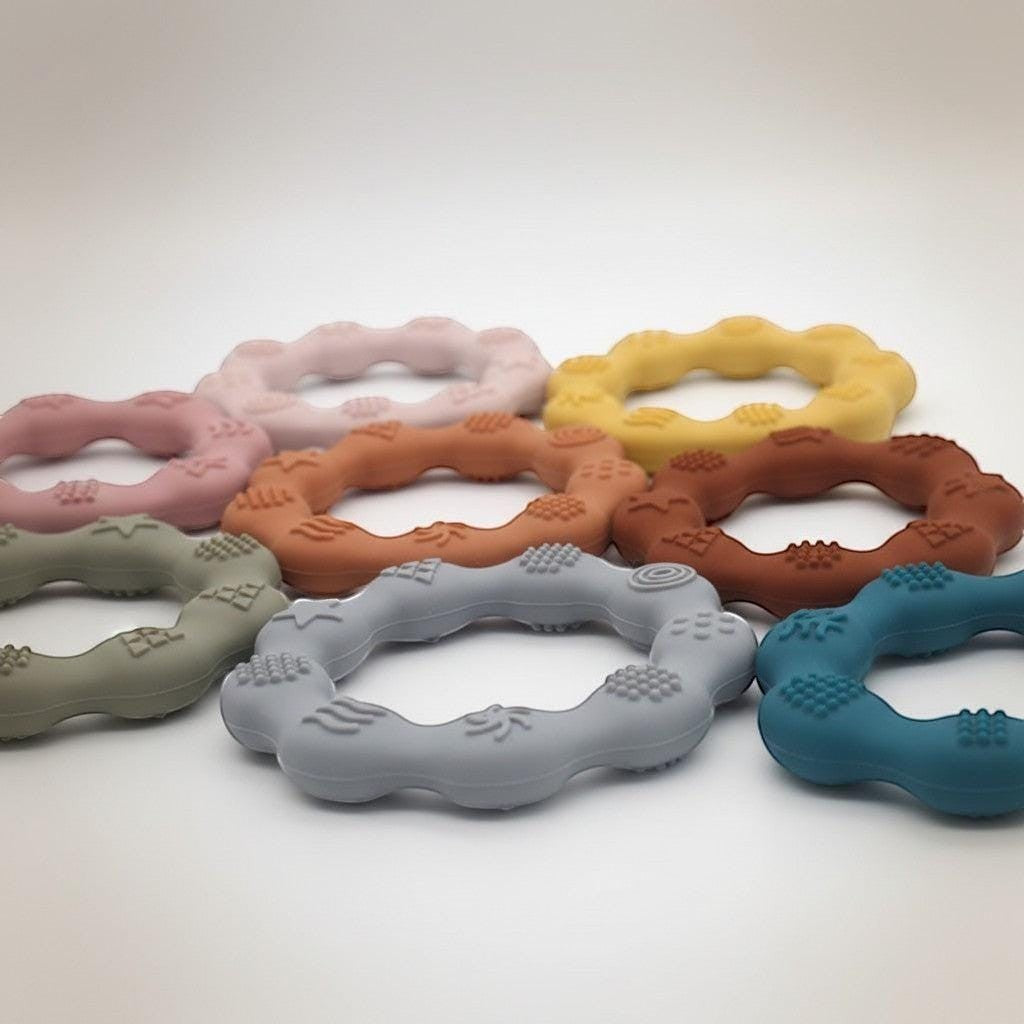 Precious Days Gifts set of 7 silicone baby teethers in pastel pink, terracotta, mustard yellow, grey, and teal with textured patterns.