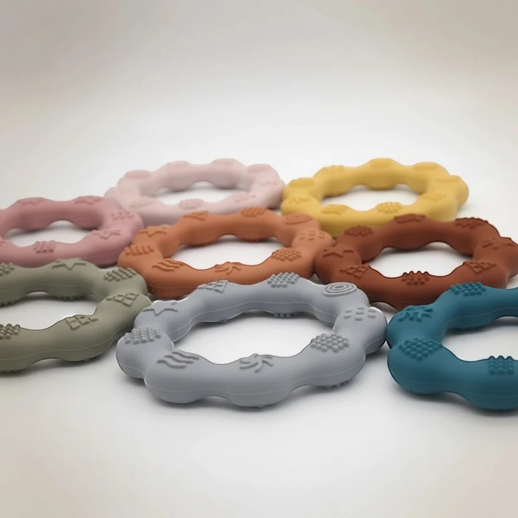 Precious Days Gifts set of silicone teethers in various pastel colors including pink, yellow, grey, and teal. Textured for baby's gums.