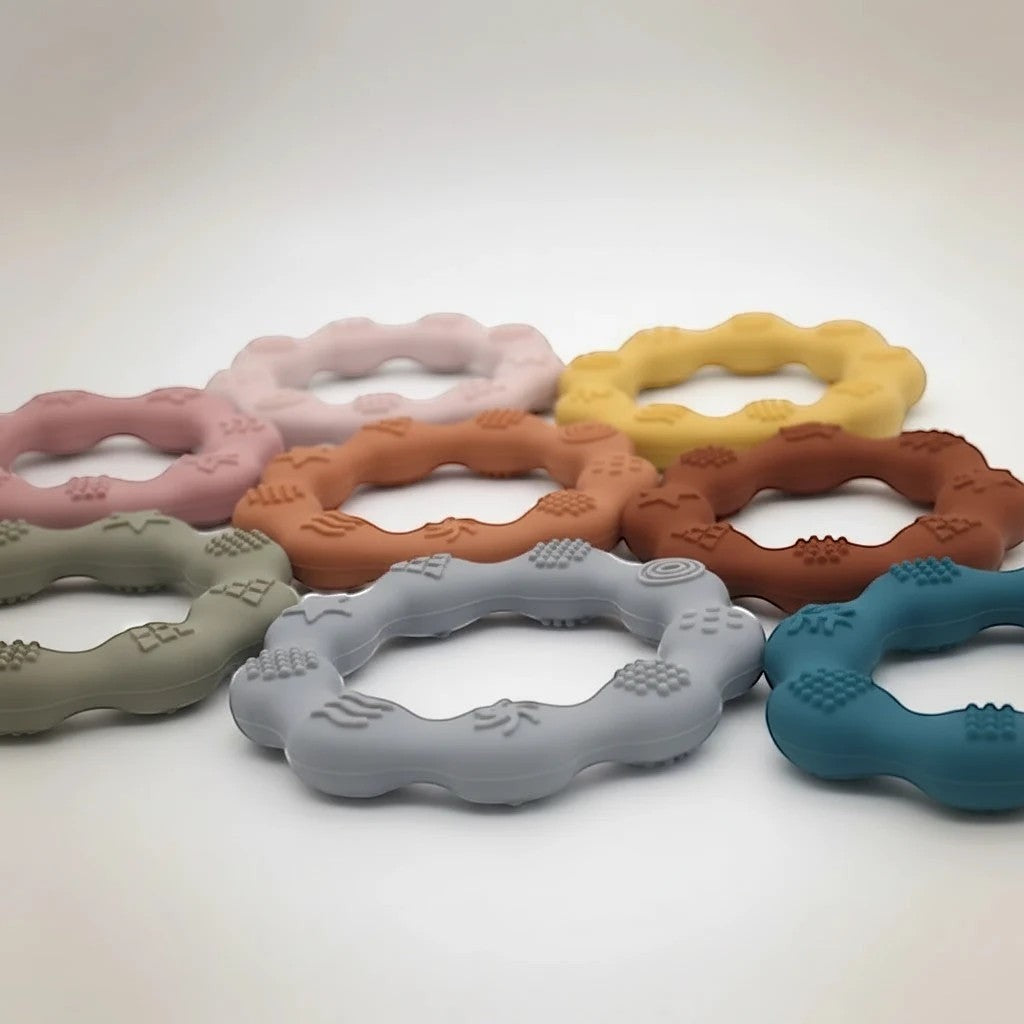 Precious Days Gifts set of silicone teethers in various pastel colors including pink, yellow, grey, and teal. Textured for baby's gums.