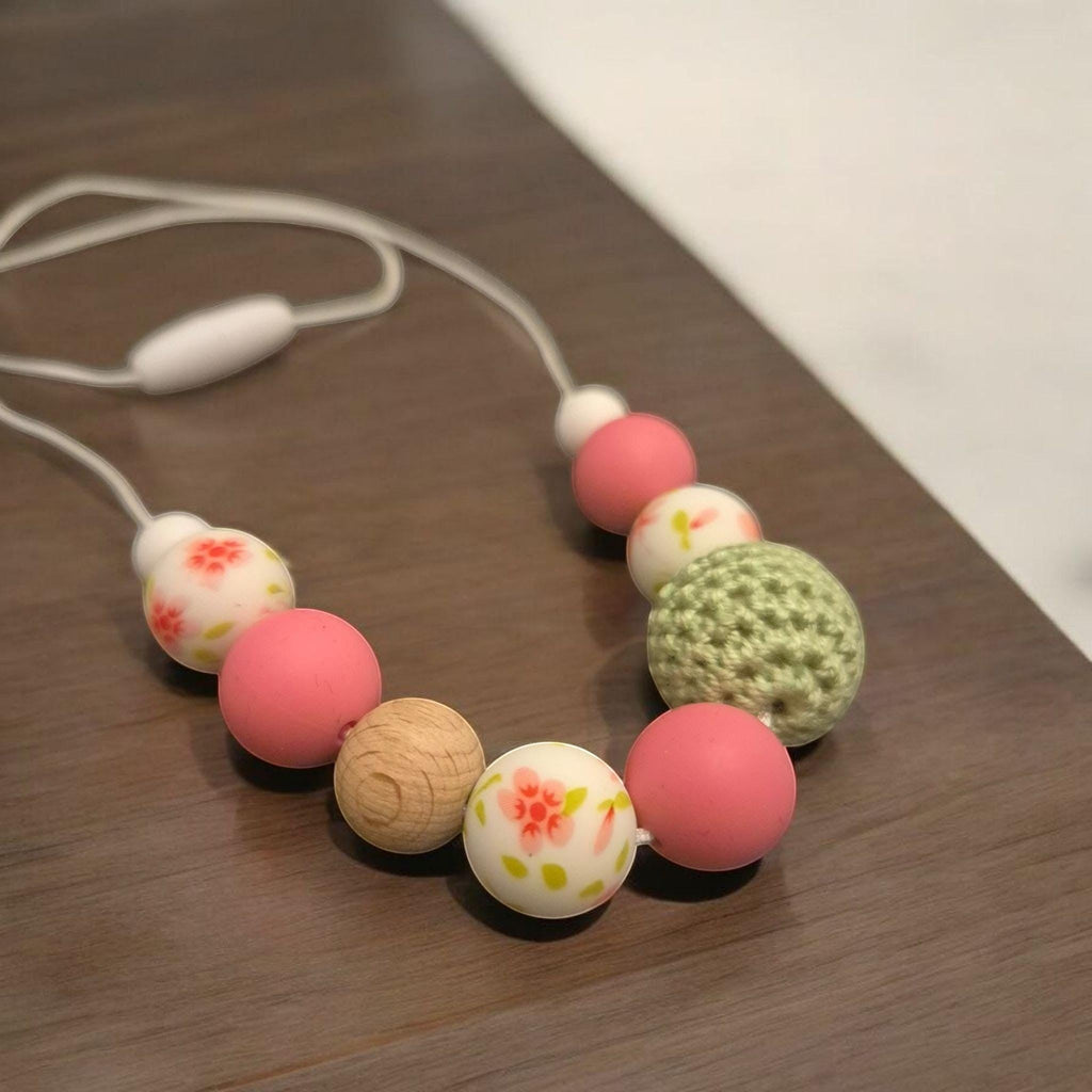 Precious Days Gifts Springtime teething necklace with pink, white floral, wood, and green crochet beads on a white cord.