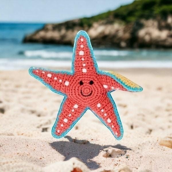 Precious Days Gifts coral starfish rattle with blue trim and white dots, smiling face, on a sandy beach.